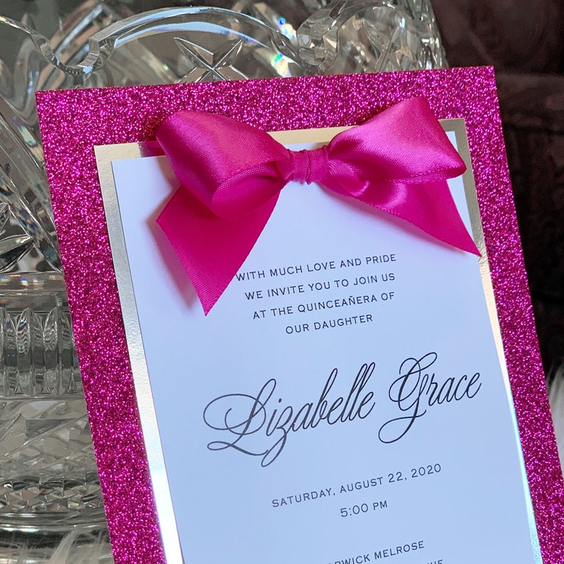May include: Pink glitter card with a gold border and a large pink bow. The card is an invitation to a Quincea&ntilde;era for Lizabelle Grace on Saturday, August 22, 2020 at 5:00 PM.
