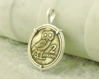 Puede incluir: Un colgante de plata con una moneda griega de dos dracmas con un diseño de búho. La moneda está engastada en un marco de plata y tiene un acabado de pátina.