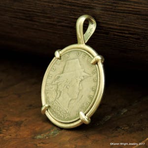 Puede incluir: Colgante de plata con una moneda antigua que muestra el perfil de una persona con sombrero. La moneda está engastada en un bisel dorado con un lazo para una cadena. El colgante se muestra sobre una superficie de madera.