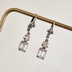 May include: A pair of silver dangle earrings with clear and iridescent crystal stones. The earrings are hanging from a gold metal stand.