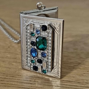 May include: A silver locket necklace with a rectangular frame and a hinged lid. The locket is decorated with a variety of colorful gemstones, including green, blue, and clear stones.