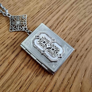 May include: A silver locket necklace with a detailed filigree design. The locket is shaped like a book and has a small diamond-shaped stone in the center.