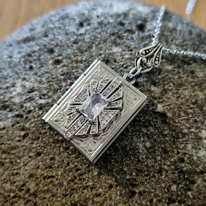 Op de afbeelding: Een zilveren ketting met een hanger in de vorm van een boek. De hanger heeft een helder kristal in het midden en een ingewikkeld ontwerp.