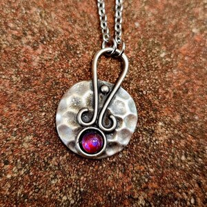Könnte beinhalten: Eine silberne Halskette mit einem gehämmerten runden Anhänger, der mit einem kleinen, schillernden rosa Edelstein verziert ist. Der Anhänger hat ein dekoratives Drahtdesign.