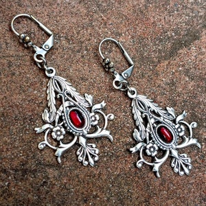 May include: A pair of silver earrings with red gemstone accents. The earrings feature a delicate floral design with intricate details.