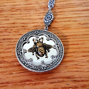 May include: A silver chain necklace with a round pendant featuring a detailed silver filigree design. The pendant has a clear glass centre with a gold bee illustration.
