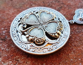 Puede incluir: Collar plateado con un colgante circular con un diseño de trébol de cuatro hojas. El colgante tiene un borde ornamentado y detallado y un elemento decorativo más pequeño en la cadena. El trébol está acentuado con detalles dorados.