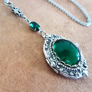 May include: A silver locket necklace with a large, oval-shaped green gemstone. The locket has a detailed, ornate design and a hinged opening.