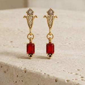 May include: A pair of gold-toned drop earrings featuring an ornate, elongated top section embellished with small clear stones. A jump ring connects the top to a rectangular, deep red faceted gemstone pendant that hangs vertically from each earring.