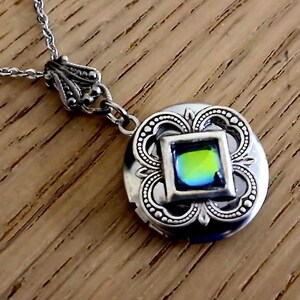 May include: A silver locket necklace with a round pendant featuring an ornate design and a square iridescent green gem in the center.