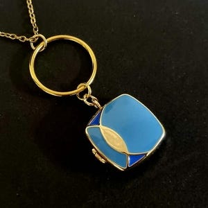 May include: A gold-tone chain necklace featuring a circular pendant loop attached to a square-shaped charm. The charm is finished with light blue enamel and features a stylised gold-tone leaf or petal design on its front surface.