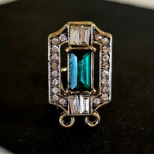 May include: A gold-toned metal jewelry component featuring a central rectangular emerald green stone, surrounded by a border of small clear crystals and two larger rectangular clear stones at the top and bottom, with two small loops at the base.