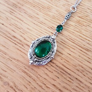 May include: A silver necklace with a large green oval gemstone pendant set in an ornate frame. The pendant is attached to a delicate silver chain with a smaller green gemstone accent.