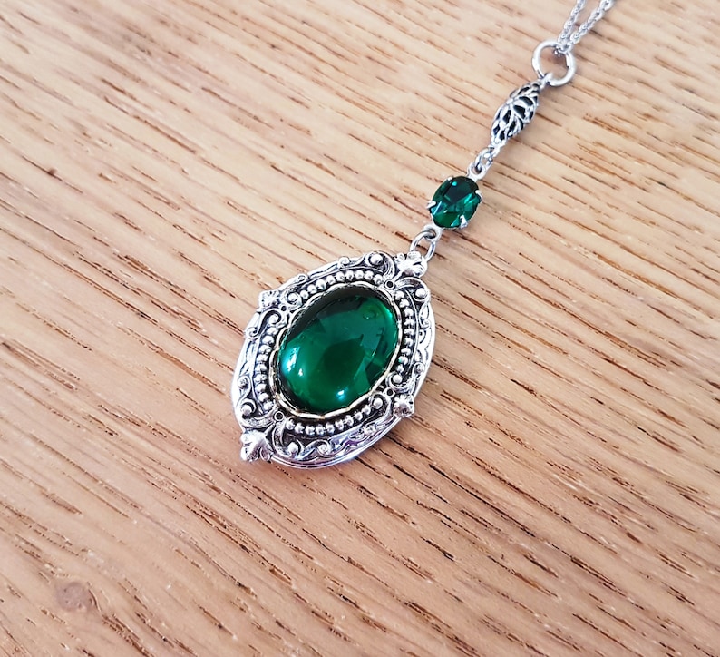May include: A silver necklace with a large green oval gemstone pendant set in an ornate frame. The pendant is attached to a delicate silver chain with a smaller green gemstone accent.