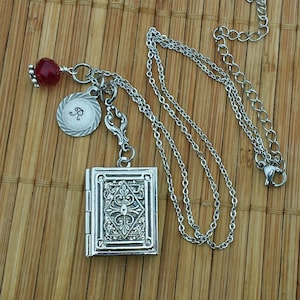 May include: A silver necklace with a book-shaped locket, a charm with the letter "R" and a red glass bead. The necklace is on a wooden surface.