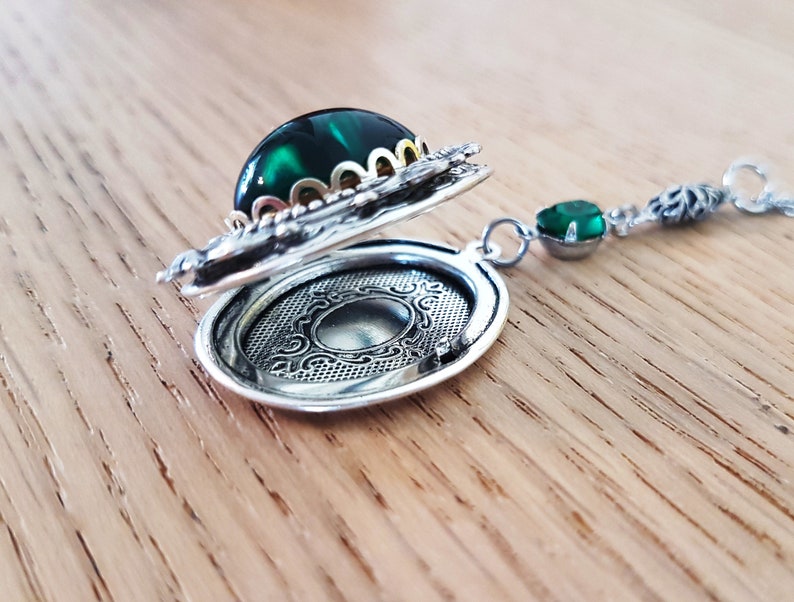 May include: A silver locket necklace with a green gemstone and ornate detailing. The locket is open, revealing a blank interior.