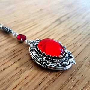 May include: A silver locket necklace with a red gemstone pendant. The locket has an ornate design and a small red crystal charm.