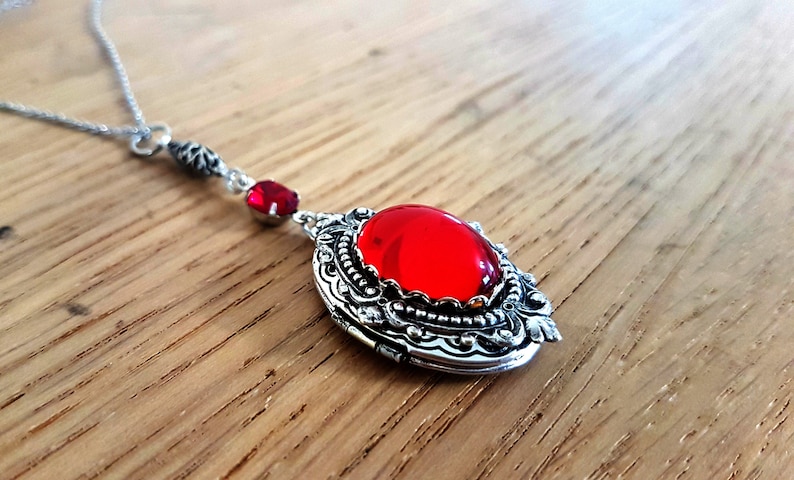 May include: A silver locket necklace with a red gemstone pendant. The locket has an ornate design and a small red crystal charm.