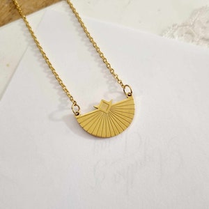 May include: A gold necklace with a geometric sunburst pendant. The pendant is a half-circle with a square in the center.