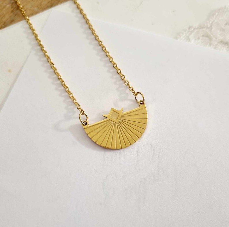 May include: A gold necklace with a geometric sunburst pendant. The pendant is a half-circle with a square in the center.