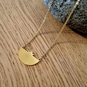 May include: A gold necklace with a geometric sunburst pendant. The pendant is a half-circle with a square in the center and radiating lines.