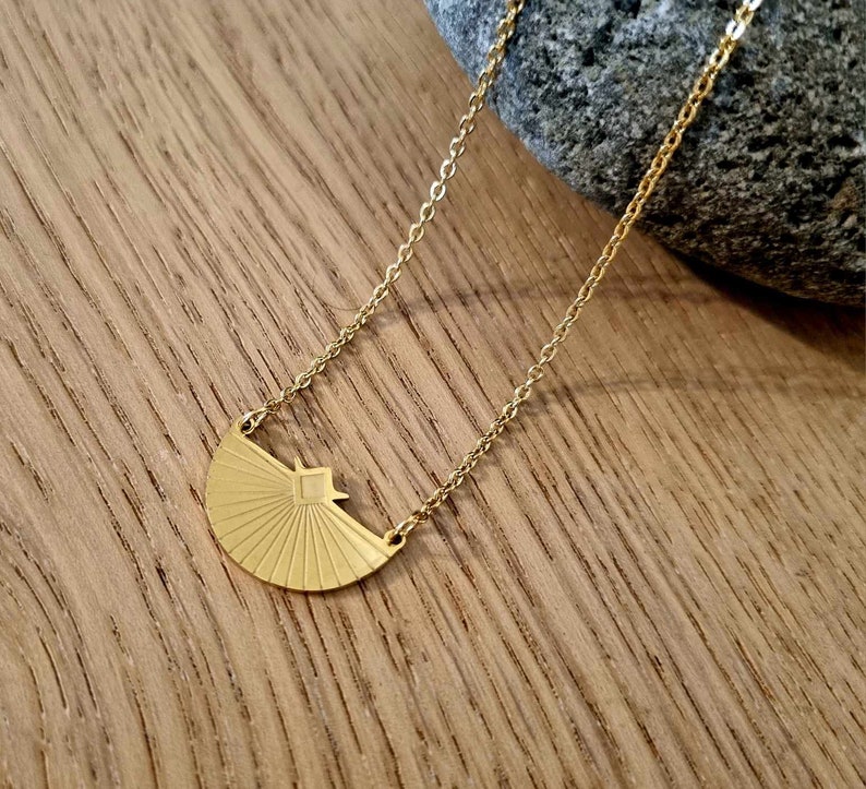 May include: A gold necklace with a geometric sunburst pendant. The pendant is a half-circle with a square in the center and radiating lines.