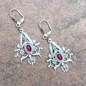 May include: A pair of silver dangle earrings with red gemstone accents. The earrings feature a floral design with intricate details.