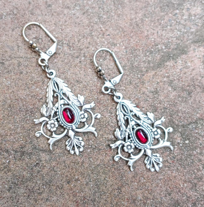 May include: A pair of silver dangle earrings with red gemstone accents. The earrings feature a floral design with intricate details.