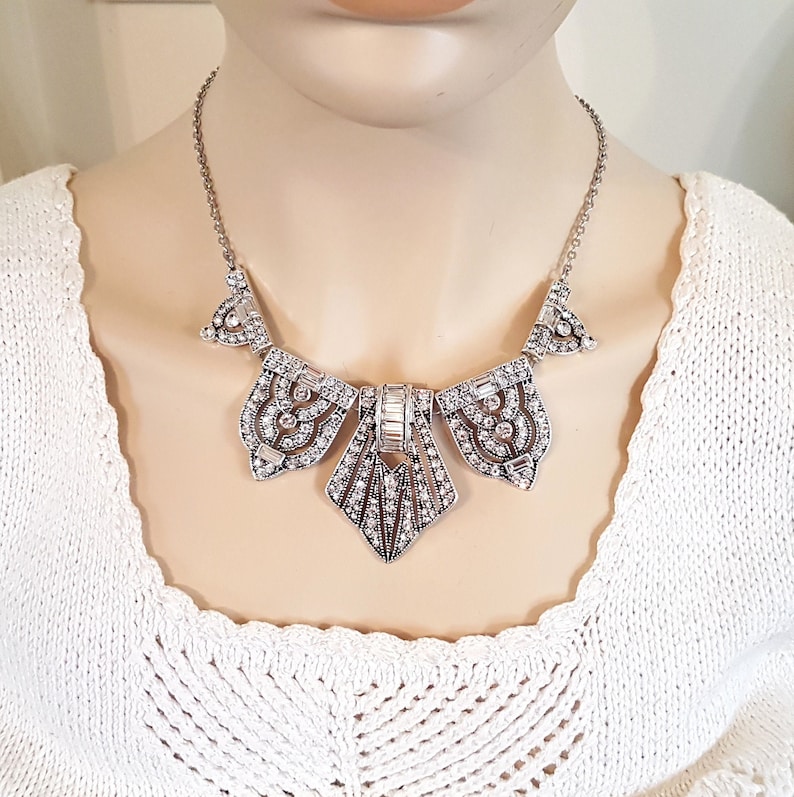 Puede incluir: Un collar de declaraci&oacute;n plateado y cristal con un dise&ntilde;o geom&eacute;trico. El collar presenta un gran colgante central con un dise&ntilde;o en forma de abanico y colgantes m&aacute;s peque&ntilde;os a cada lado.