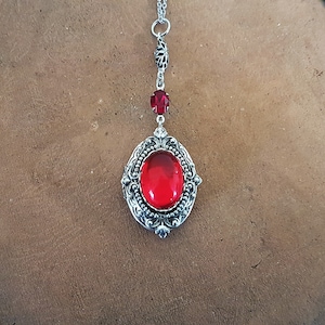 May include: A silver locket necklace with a red gemstone. The locket has an ornate design and is attached to a silver chain.
