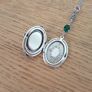 May include: Silver locket necklace with an oval-shaped pendant. The locket is open, revealing a blank interior. The necklace features a green crystal charm.
