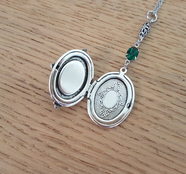 May include: Silver locket necklace with an oval-shaped pendant. The locket is open, revealing a blank interior. The necklace features a green crystal charm.
