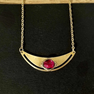 Op de afbeelding: Een gouden ketting met een halve maan hanger met een rode edelsteen. De hanger heeft een delicaat ontwerp met een klein, versierd frame rond de edelsteen.