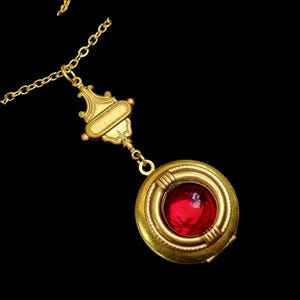 Pode incluir: Um colar com pingente dourado com um medalhão redondo com uma grande pedra preciosa vermelha. O medalhão é suspenso por uma corrente dourada decorativa e um conector ornamentado. O fundo é preto.