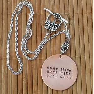 Pode incluir: Um colar com pingente de cor de cobre com as palavras "ever thine ever mine ever ours" estampadas no pingente redondo. O colar tem uma corrente prateada e um fecho decorativo com detalhes florais. O colar &eacute; exibido em uma superf&iacute;cie de bambu.