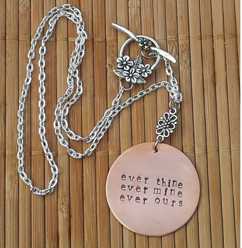 Pode incluir: Um colar com pingente de cor de cobre com as palavras "ever thine ever mine ever ours" estampadas no pingente redondo. O colar tem uma corrente prateada e um fecho decorativo com detalhes florais. O colar &eacute; exibido em uma superf&iacute;cie de bambu.