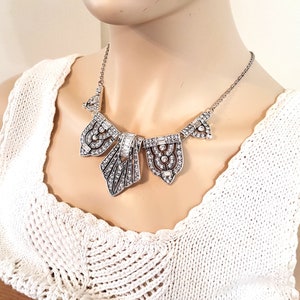 Puede incluir: Un collar de declaraci&oacute;n plateado con un dise&ntilde;o geom&eacute;trico que presenta m&uacute;ltiples colgantes incrustados con cristales. El collar est&aacute; en un maniqu&iacute; que lleva una blusa de crochet blanca.
