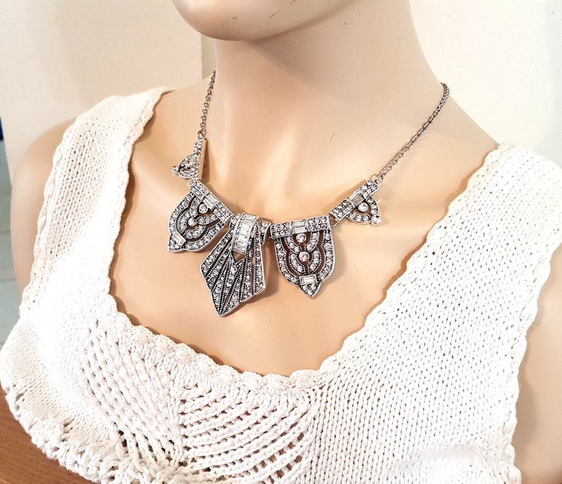 Puede incluir: Un collar de declaraci&oacute;n plateado con un dise&ntilde;o geom&eacute;trico que presenta m&uacute;ltiples colgantes incrustados con cristales. El collar est&aacute; en un maniqu&iacute; que lleva una blusa de crochet blanca.