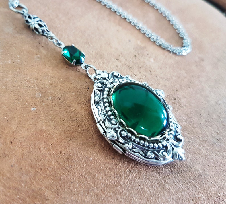 May include: A silver locket necklace with a large, oval-shaped green gemstone. The locket has a detailed, ornate design and a hinged opening.