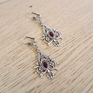 May include: Silver dangle earrings with a red gemstone and intricate floral designs.
