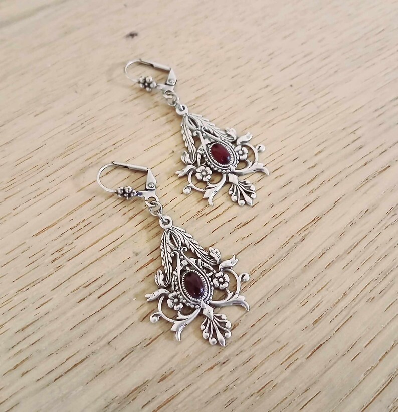 May include: Silver dangle earrings with a red gemstone and intricate floral designs.