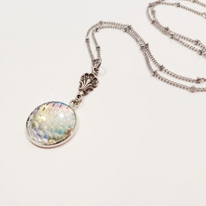 May include: A silver chain necklace with a round iridescent pendant featuring a mermaid scale design. The pendant is attached to a silver charm with a floral design.
