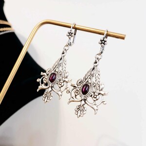 May include: Silver dangle earrings with a floral design and a dark purple gemstone in the center.
