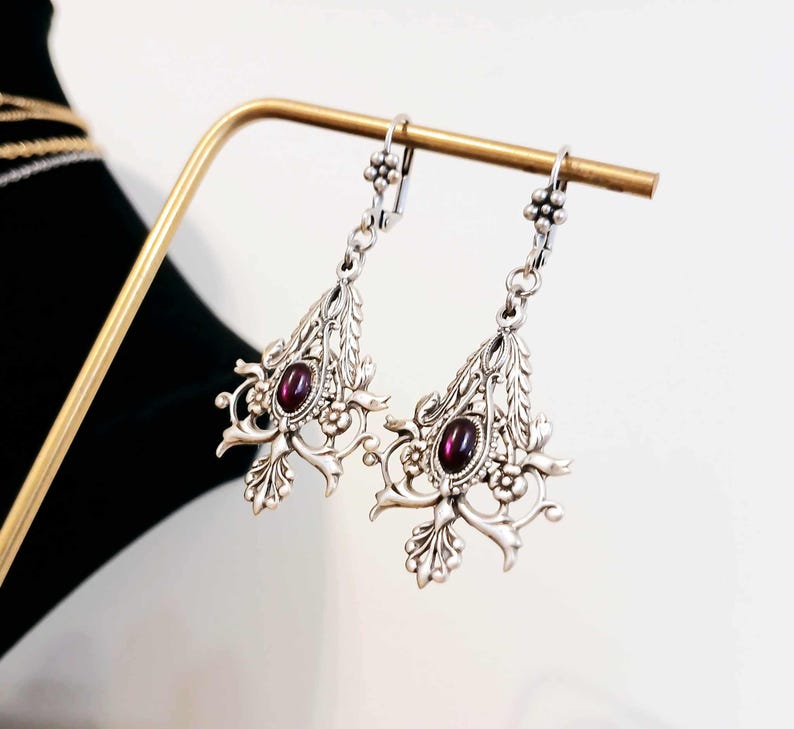 May include: Silver dangle earrings with a floral design and a dark purple gemstone in the center.