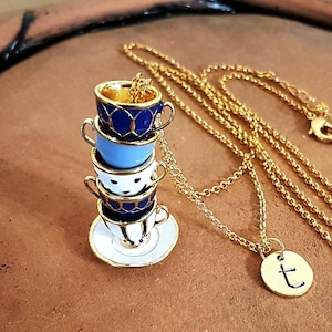 Op de afbeelding: Een gouden ketting met een bedel van een stapel van vier miniatuur theekopjes met blauwe en witte patronen. De bedel staat op een wit schoteltje. De ketting heeft ook een kleine gouden bedel met de letter 't' erop.