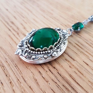 May include: A silver locket with an ornate design and a large, green gemstone. The locket is on a chain with a smaller green gemstone.