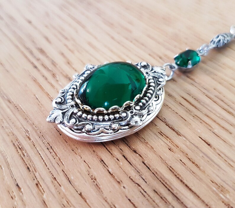 May include: A silver locket with an ornate design and a large, green gemstone. The locket is on a chain with a smaller green gemstone.