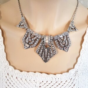 Puede incluir: Un collar de declaraci&oacute;n plateado con un dise&ntilde;o geom&eacute;trico adornado con diamantes de imitaci&oacute;n transparentes. El collar tiene una cadena delicada y un gran colgante adornado.