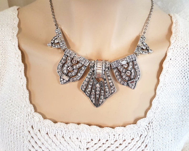 Puede incluir: Un collar de declaraci&oacute;n plateado con un dise&ntilde;o geom&eacute;trico adornado con diamantes de imitaci&oacute;n transparentes. El collar tiene una cadena delicada y un gran colgante adornado.