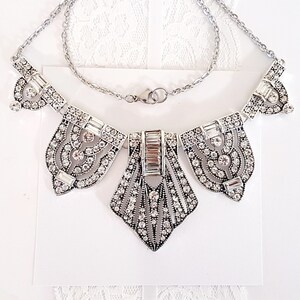 Puede incluir: Un collar de plata con un dise&ntilde;o geom&eacute;trico que presenta un colgante central en forma de diamante y dos colgantes m&aacute;s peque&ntilde;os a cada lado. El collar est&aacute; adornado con diamantes de imitaci&oacute;n transparentes.